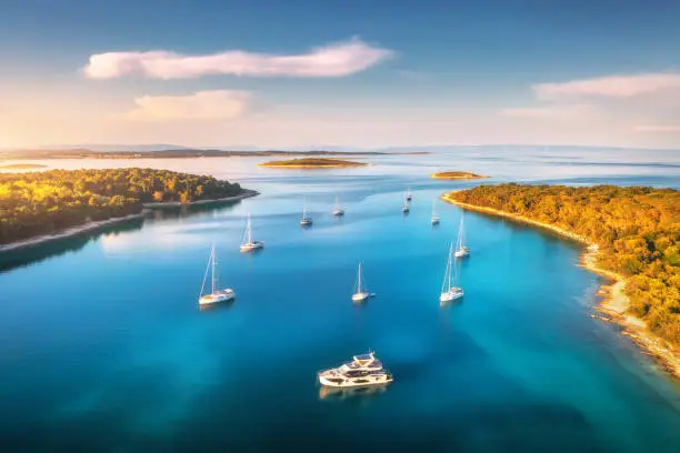 Photo of Aerial view of beautiful yachts and boats on the sea at sunset in summer. Adriatic sea, Kamenjak, Croatia. Top view of luxury yachts, sailboats, lagoon, clear blue water, and green forest. Travel