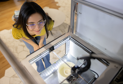 Happy Latin American woman working at a design studio and watching a 3D printer while printing a model - high angle view