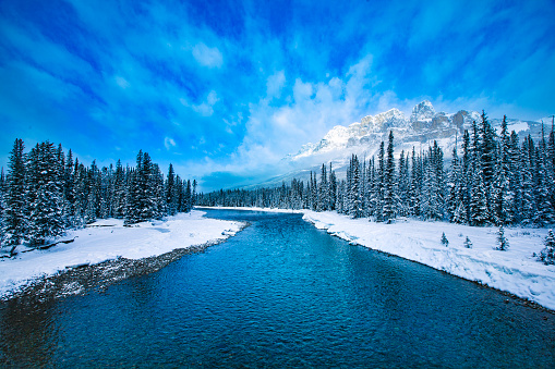 Idyllic winter scene of bright blue snowmelt river, snow covered mountain landscape and bright blue sky with dramatic cloud formation