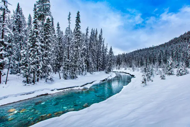 Photo of Idyllic winter scene of bright blue snowmelt river, snow covered mountain landscape and bright blue sky with dramatic cloud formation