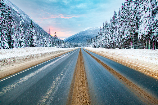 Winter road trip scene of snow covered road lined with pine trees and dramatic sunset