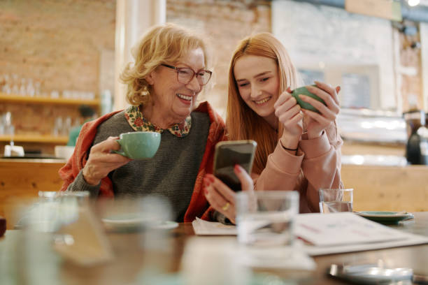 Nonna felice e sua nipote adolescente seduti in un bar, gustando un caffè e usando lo smartphone per video divertenti. - foto stock