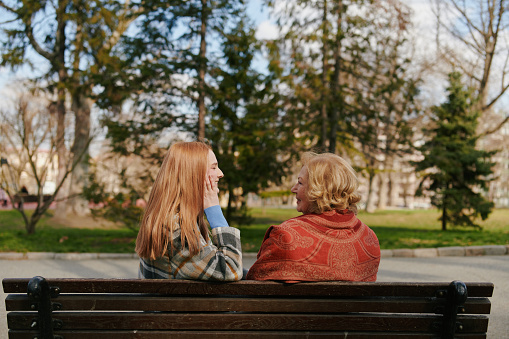 A young ginger woman is sitting with her grandmother and talking on the park bench in the cold weather.