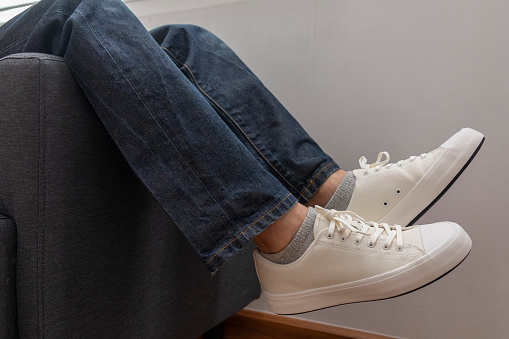 Close up of man resting and relaxing feet in cozy comfortable living room. White sneaker and blue jean with sofa.