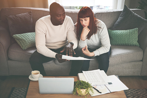 A couple going through paperwork at home and using a laptop. A young man and woman paying bills, planning their finance and looking at paper online with a computer on the sofa