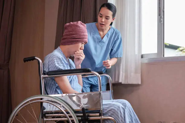 Uniformed young Asian female therapy doctor with wheelchair male patient at window to support recovery, suffer and cough from cancer illness after chemo medical treatment in hospital inpatient room.