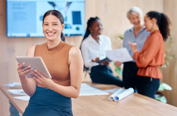 istock Beautiful young mixed race business woman using a tablet while leaning against the boardroom table after a meeting with her female only colleagues in the background. Our office is going digital 1398739977