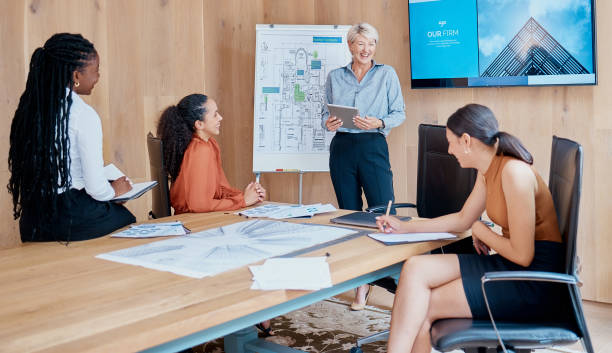istock Mature caucasian businesswoman smiling and standing using a tablet while giving a presentation in the boardroom during a meeting with her female only diverse multiracial colleagues in a workplace. Our office is going digital 1398740194