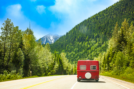 Road trip scene with vintage red caravan driving through rich green pine forest mountains and blue sky