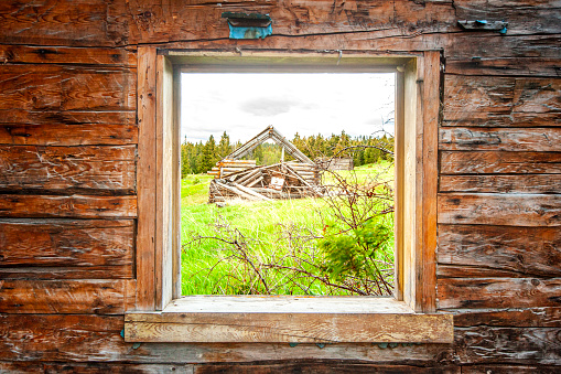 Old wooden window shutter in authentic log cabin. Visible inside are wooden bed and animal scull hanging in rafters. Light color chink fills spaces between weathered logs. Part of building in Fort William Historical Park, Thunder Bay, Ontario.