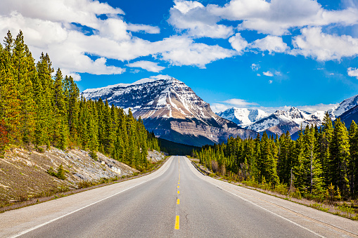 Road trip scene with straight open road, blue sky, snow capped mountains, valleys and pine tree forests