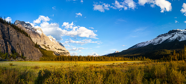 Snow capped rocky mountain terrain with blue sky and clouds with flat meadow in warm golden light