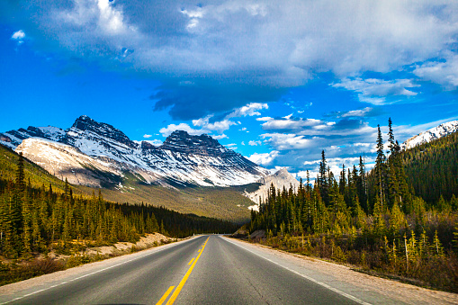 Road trip scene with straight open road, blue sky, snow capped mountains, valleys and pine tree forests