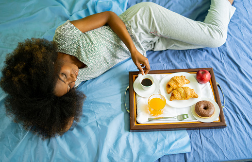 A woman enjoying her breakfast in bed of croissant, apple, orange juice, hot black coffee and a cheeky chocolate donut, while she is lying on the bed, viewed from above