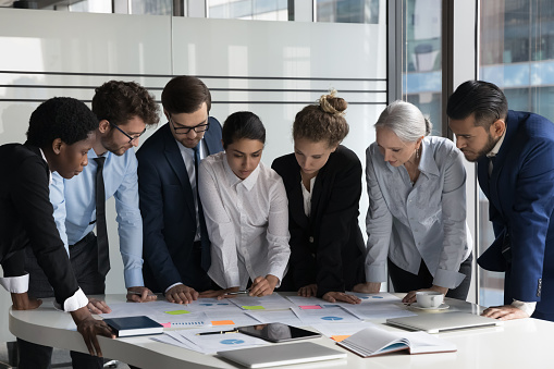 Multiethnic business team meeting over project marketing reports, analyzing paper charts with sticky notes, using scrum method, working on strategy, business plan. Diversity, teamwork concept