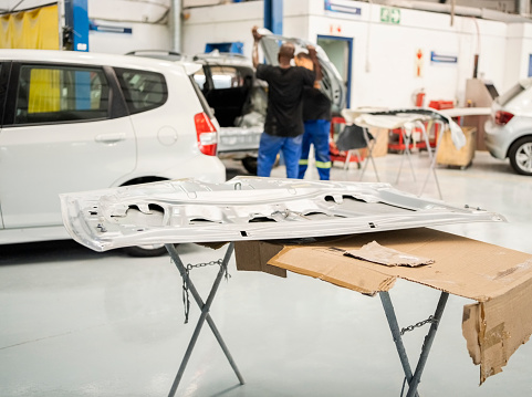 Vehicle bonnet hood on table in front with two mechanics working in background at automobile repairing workshop