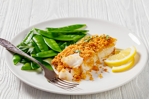 Baked Haddock with Crackers butter toppings served with boiled snow peas and lemon on white plate on white wood table