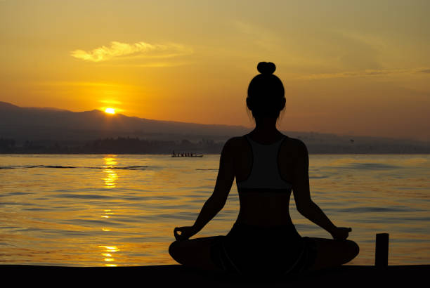 Woman silhouette in Yoga Easy Meditating pose at sunrise with very quiet ocean Woman silhouette in Yoga Easy Meditating pose at sunrise with very quiet ocean in background sukhasana stock pictures, royalty-free photos & images