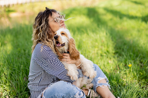 Beautiful young woman sitting on green grass and hugging her white Cocker Spaniel puppy on sunny spring day