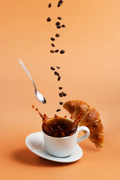 White coffee cup falling on beige table background, splashes, beans, croissant