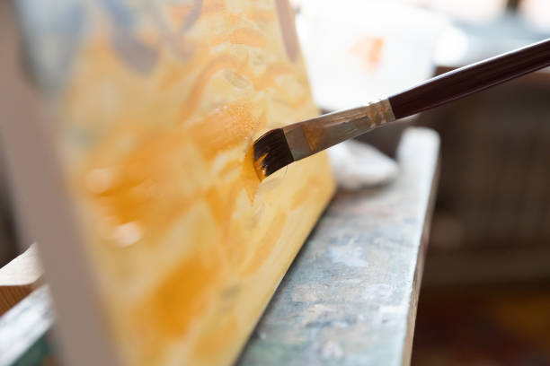 Close up of paintbrush bristle touching canvas. Artist painting picture, applying yellow strokes on canvas, working on artwork details at easel. Cropped shot