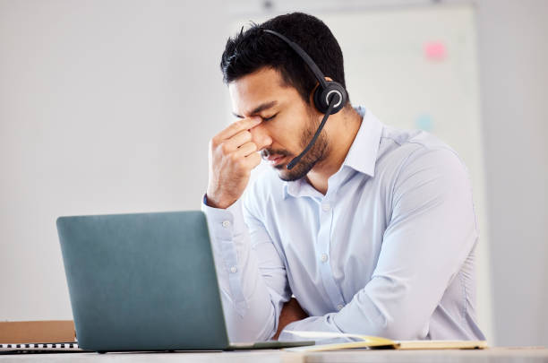 giovane uomo di razza mista che sembra stressato mentre lavora come agente di call center. uomo bello che soffre di depressione e mal di testa mentre indossa un auricolare e lavora in un lavoro d'ufficio - frustration telephone computer it support foto e immagini stock