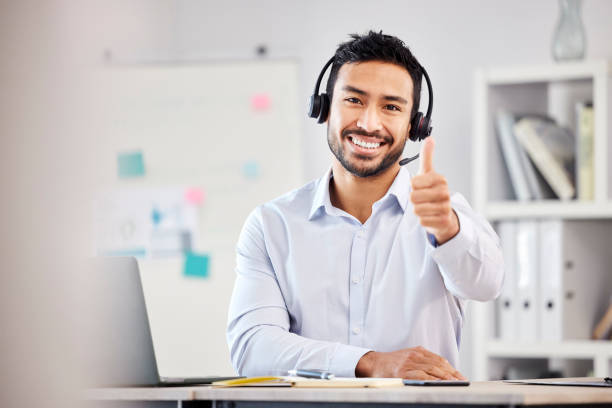 jovem feliz e bonito agente de call center masculino mestiço mostrando um polegar para cima usando um fone de ouvido sentado em uma mesa em um escritório sozinho no trabalho. satisfeito agente hispânico de atendimento ao cliente atendendo chamadas - looking at camera smiling desk isolated - fotografias e filmes do acervo