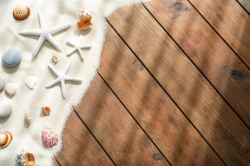 Summer vacations and marine background: starfish, conch shells, and sea stones on tropical white sand border shot from above on wooden plank. The composition is at the left of an horizontal frame leaving useful copy space for text and/or logo at the right. High resolution 42Mp digital capture taken with SONY A7rII and Zeiss Batis 40mm F2.0 CF lens