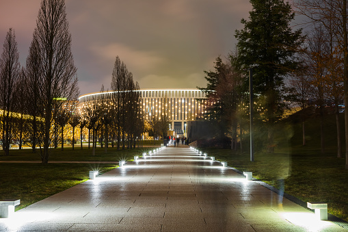 Evening park with a pedestrian illuminated path leading to semicircular large luminous building. Blurred image. Motion blur