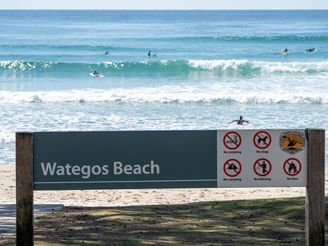 Horizontal closeup photo of the painted wooden sign for 'Wategos Beach', with several 'forbidden' symbols on it, on the grass area above the beach. In the background, waves roll in and break on the sandy beach. Wategos Beach, Byron Bay, north coast NSW. August 26th, 2021.