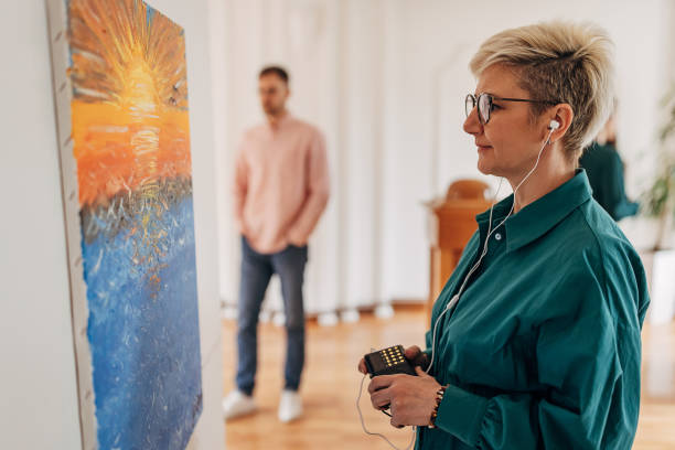 Diverse group of visitors in modern art gallery looking at paintings. woman looking at paintings and listening to audio guide on headphones.