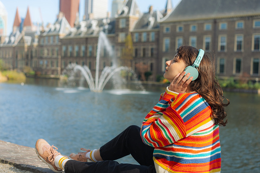 Young Caucasian woman in colourful sweater  listening to music in headphones near the lake  in The Hague