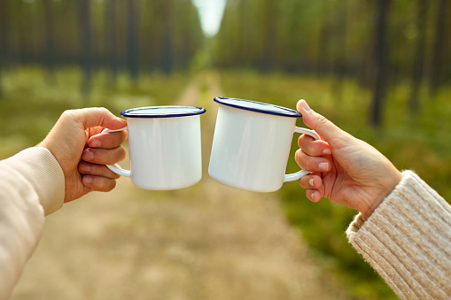picking season, leisure and people concept - hands of man and woman clinking white tin tea mugs in forest