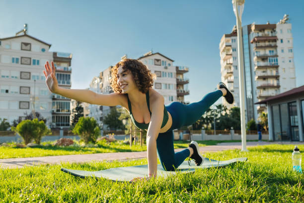 Young happy redhead woman wearing sportive clothes on yoga mat on city park, outdoors practicing bird dog pose, knee to forehead curl exercise. Concept of healthy lifestyle, yoga. stock photo