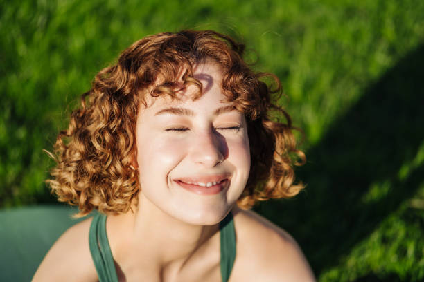Redhead young woman wearing green sports bra sitting on yoga mat on city park, outdoors closed eyes with cute expression. stock photo