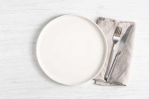 White empty plate and silver cutlery on linen napkin on white wooden table.