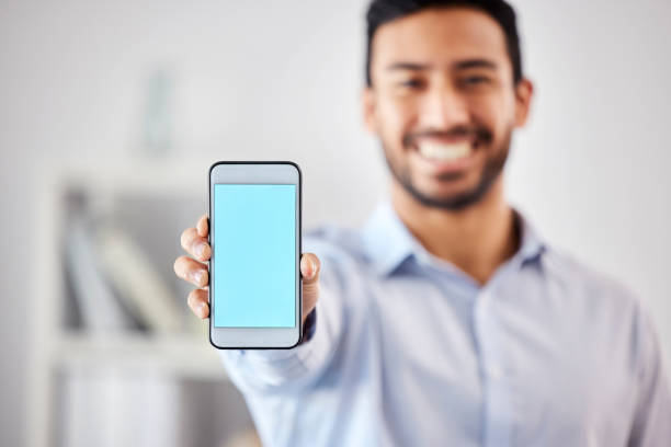 istock Businessman showing a blank blue screen with copyspace on his cellphone in the office. Mixed race professional holding his phone to display a new webpage and website. Corporate man using technology 1398810330