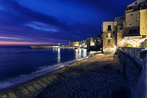 Panorama of Cefalu, Sicily harbor at night. Shot from the promenade. The city old town buildings meet the sea at a small beach.\nShot with Canon R5