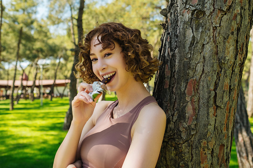 Young sporty redhead woman resting while biting a nutritive bar while leaning to tree looking at the camera on green city park, outdoor.