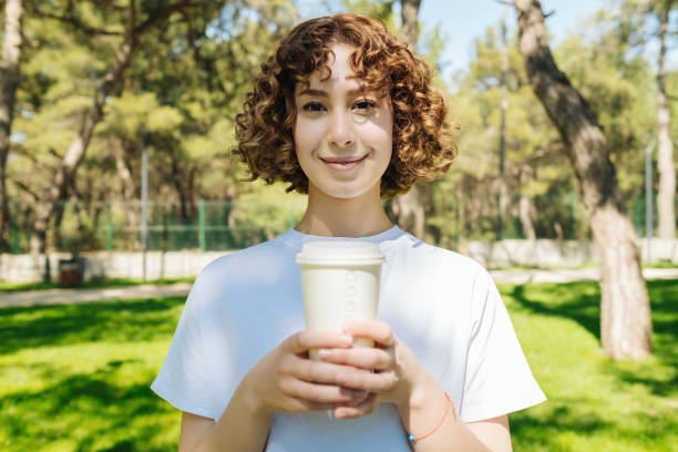 istock Happy young redhead woman wearing white tshirt holding takeaway coffee cup while standing on green city park, outdoors. 1398812254