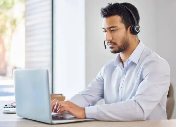 Photo of Businessman working in a call center. Operator working on his laptop. Young multiracial man working, wearing a headset. Young businessman working in customer service, on a call.