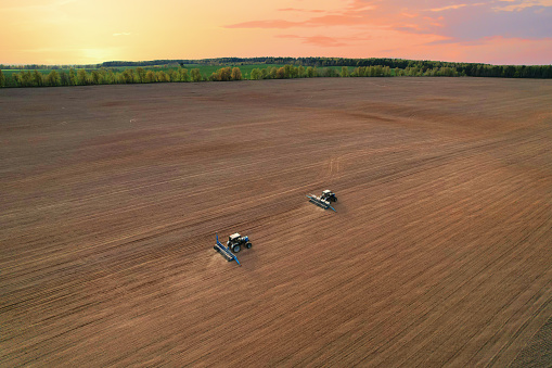 Tractor sowing seed on plowed field. Sowing seeds of corn and sunflower. Blue Tractor with disk harrow on plowing field. Seeding machinery on farm field. Seed sowing in farmland on sunset.