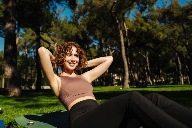 Sporty smiling redhead woman wearing sports bra and doing sit ups abs crunches exercise outdoor lying on yoga mat. Sport workout routine, morning physical training exercises. Outdoor sport concept. stock photo