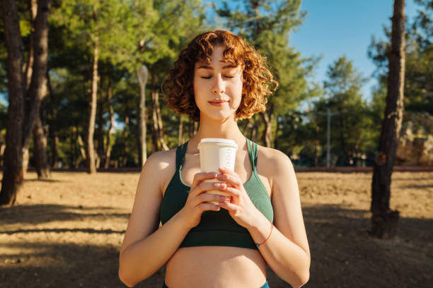 Beautiful redhead sportive woman wearing green sports bra standing on city park, outdoors holding a takeaway coffee mug front of chest and closed eyes. Coffee lover and outdoor sport concepts. stock photo