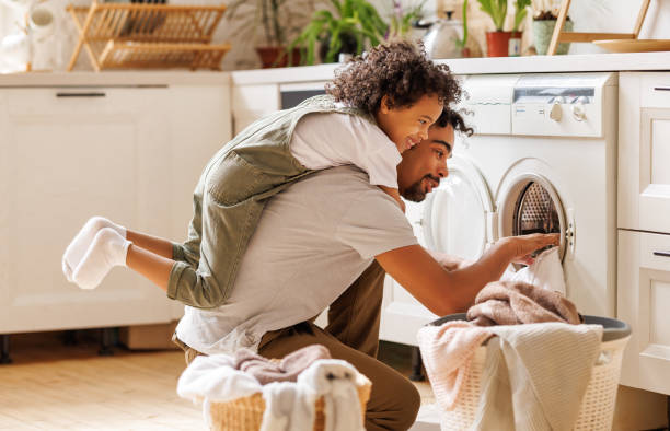 Father and son doing laundry together Side view of black child in casual clothes with curly hair smiling and embracing dad loading washing machine during household routine in morning at home washer stock pictures, royalty-free photos & images