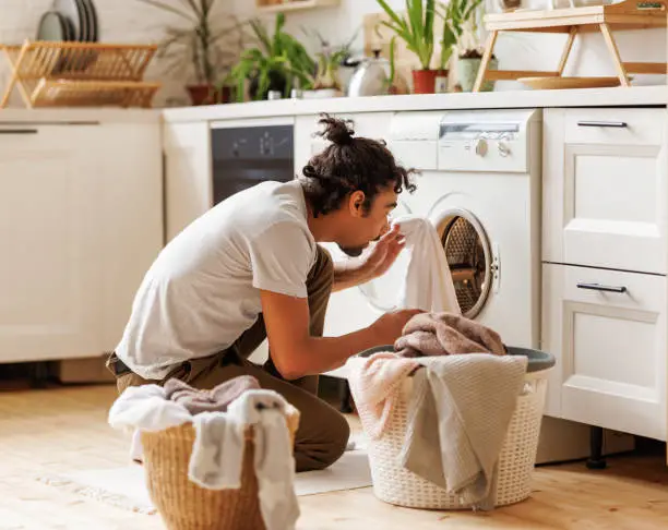 Young ethnic black man householder with curly hair sitting on floor near  washing machine while doing laundry in kitchen at home