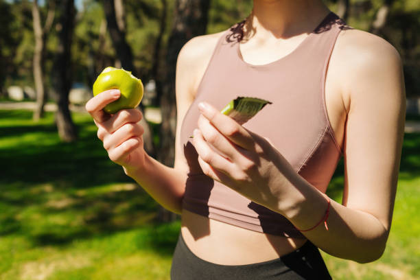 Beautiful sporty woman eating apple and holding protein bar while resting from exercise. Healthy snack for fitness girl. Fitness beautiful woman eating an energy snack outdoor. stock photo