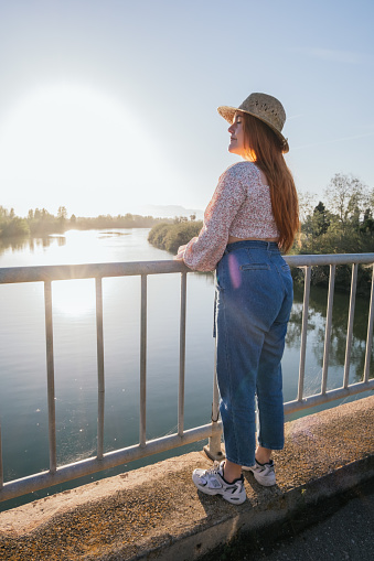 red-haired girl wearing a straw hat watching a sunset from a bridge