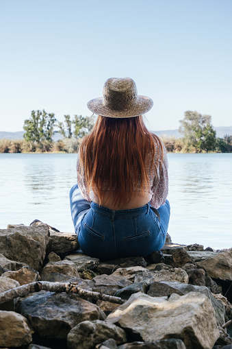 red-haired girl in a straw hat sitting on a stones looking at a river