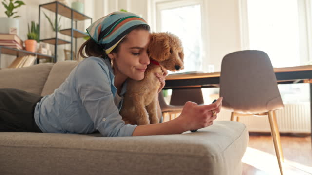 Young woman relaxing on sofa with her dog at home using smart phone